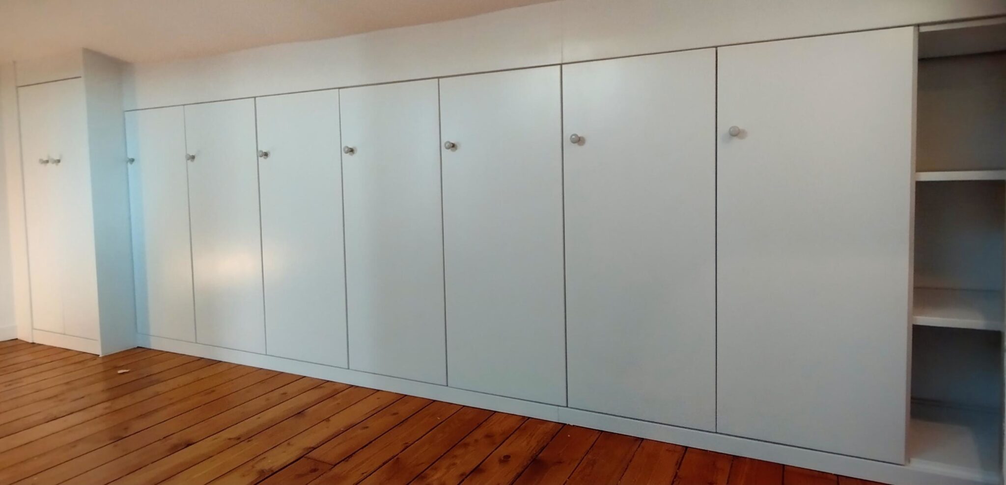 Penderie et étagères réglables intégrées dans un dressing sous pente en mélaminé blanc.